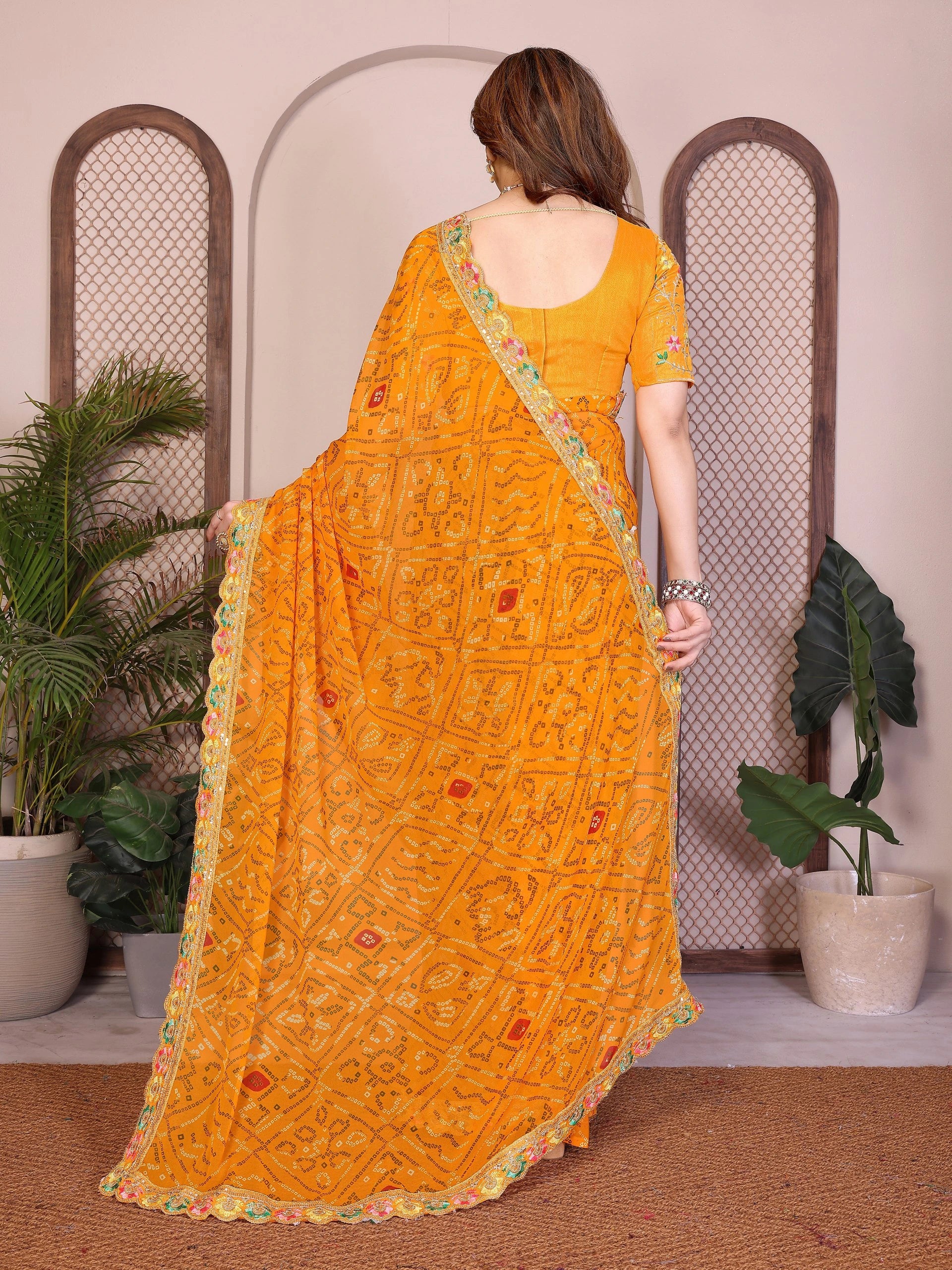 Woman wearing a yellow saree with a pattern, standing in a room with plants.